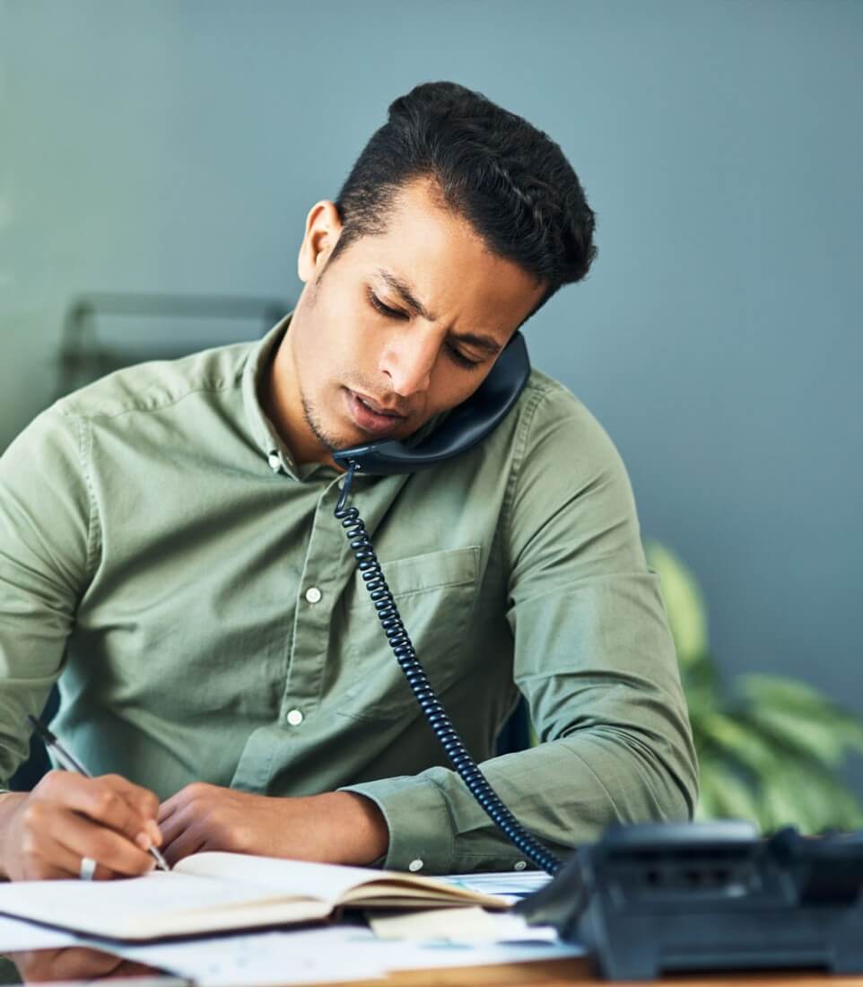 A man writing in a journal while he speaks on the phone, he has a mixed complexion, light stubble and short dark hair.