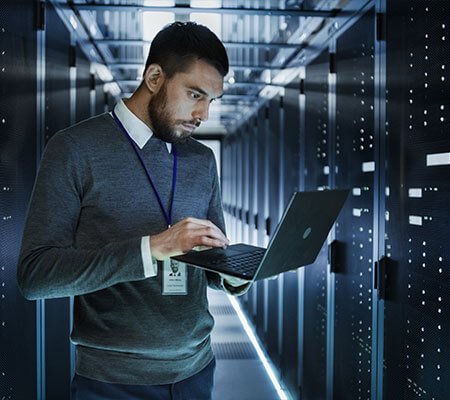 A man walking through a server room with a laptop in his hand.