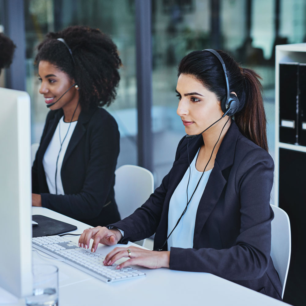 Two women sit next to each other, each with a headset on, speaking on a mic that is a part of their VoIP telephone system.